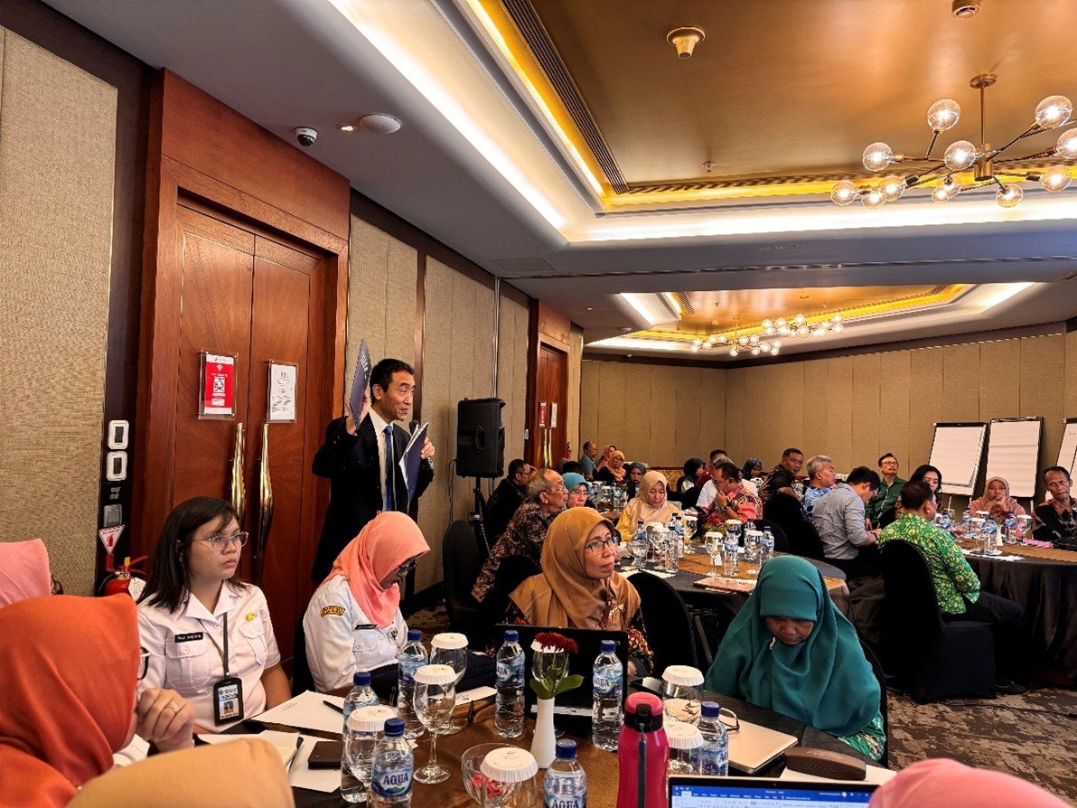 A man is speaking at a workshop, holding a booklet in his hands, while a diverse group of participants listens attentively.
